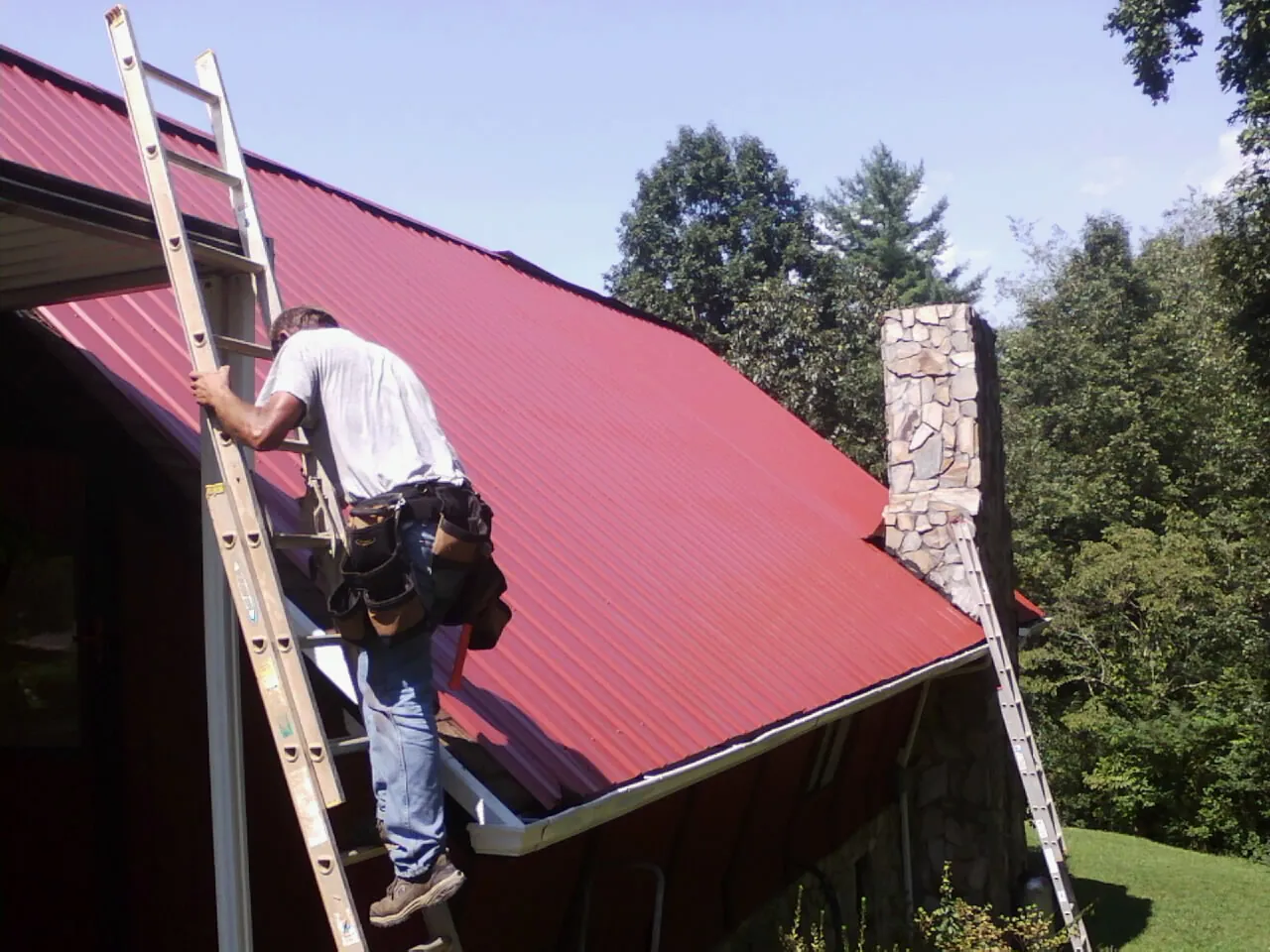 Roofer on a ladder beside a red metal roof and stone chimney — Soffit & Fascia Repair by Roofing Brothers in Redwood City